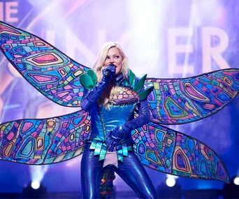 Singer in a colorful bug-themed costume performs on stage with vibrant background lights.