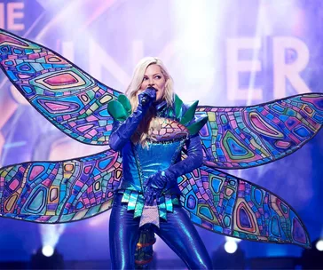 Singer in a colorful bug-themed costume performs on stage with vibrant background lights.