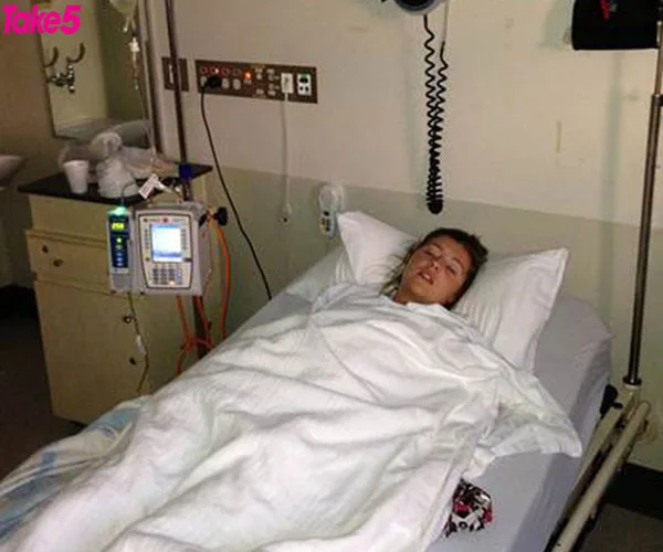 Person lying in a hospital bed, covered with a white blanket, with medical equipment nearby.