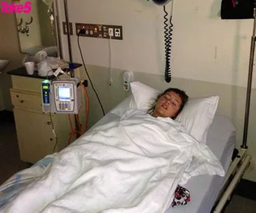 Person lying in a hospital bed, covered with a white blanket, with medical equipment nearby.