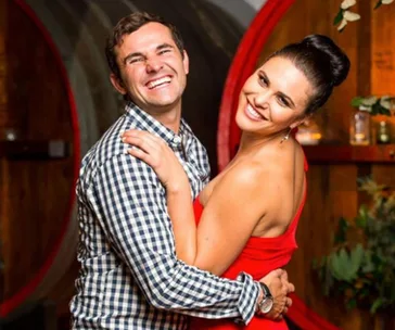 Man and woman smiling and embracing in front of large wooden barrels.