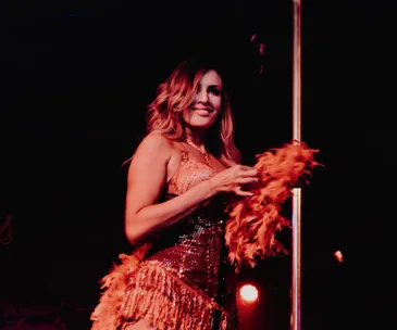 A woman in a glittery outfit performs on stage with a pole, holding an orange feather boa, smiling under stage lights.