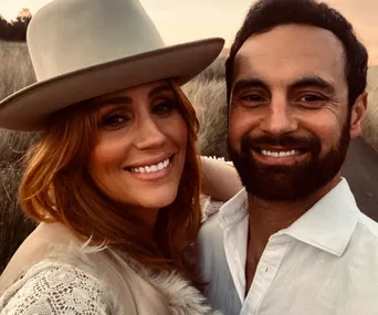 A smiling couple takes a selfie in a grassy field at sunset, with the woman wearing a wide-brimmed hat.