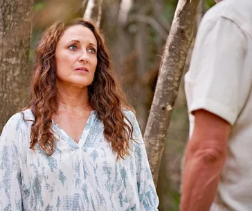 A woman with long brown hair in a blue patterned top looks at a man in the forest.