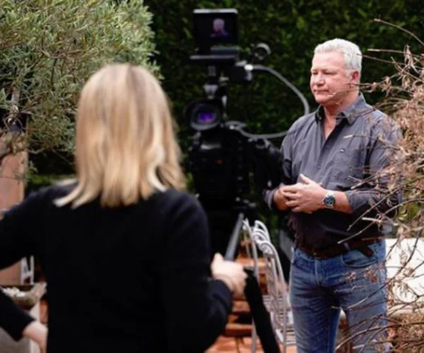 A man stands outdoors with a microphone, facing a camera, while another person is seen from behind.