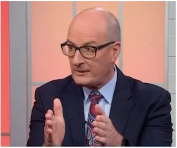 A person in a suit and glasses is speaking with hand gestures on a set with a warm background.