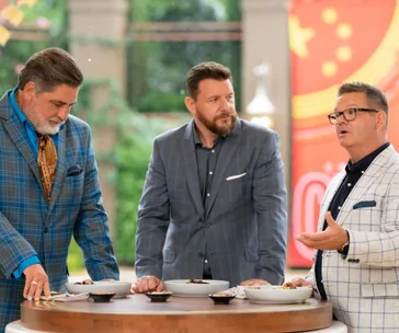 Three men in suits discuss around a table with dishes. They're judges on a cooking show.