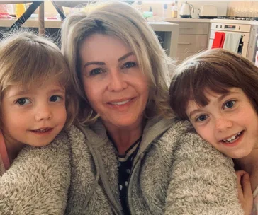 A woman smiles with two young children, one on each side, in a cozy kitchen setting.