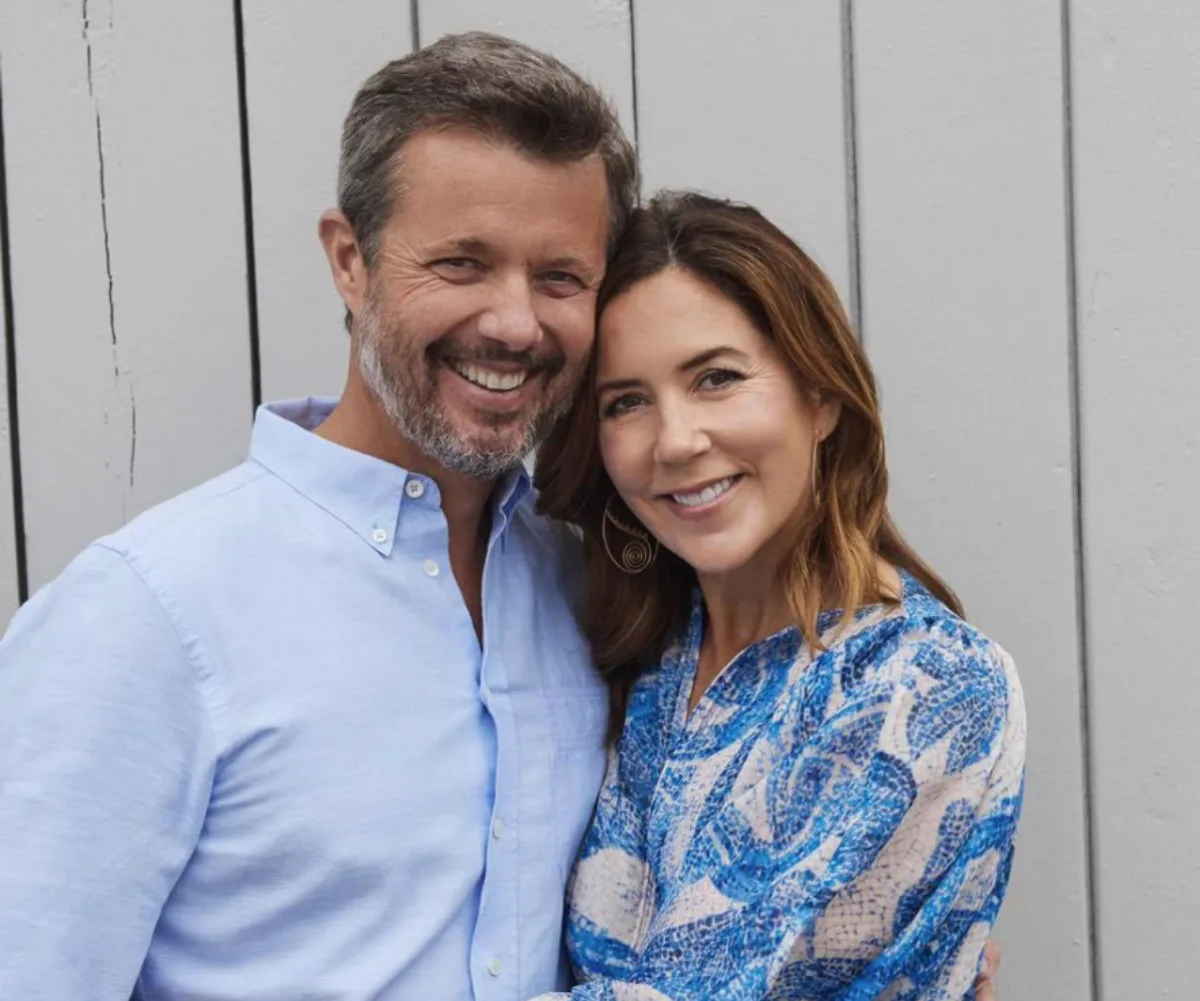 Couple smiling together, man in light blue shirt and woman in blue patterned dress, standing against a wooden wall.