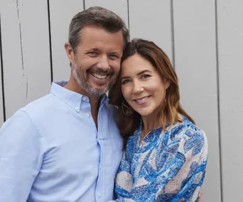 Couple smiling together, man in light blue shirt and woman in blue patterned dress, standing against a wooden wall.