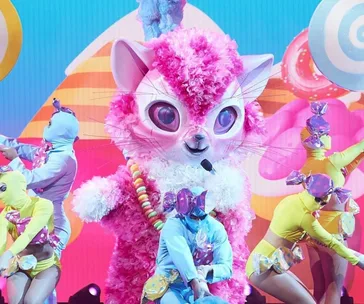 Kitten costume character with pink fur performs on stage with colorful, candy-themed background and dancers in vibrant outfits.
