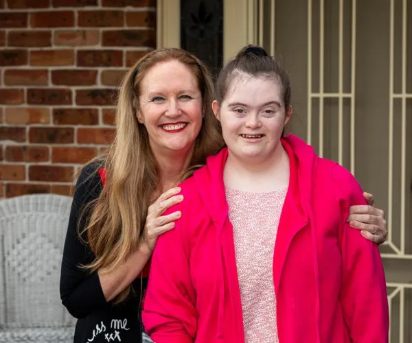Woman with red hair and black outfit embraces a young woman in a pink jacket, both smiling in front of a brick wall.