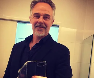 Man taking a mirror selfie in a bathroom, wearing a dark jacket and shirt, smiling with gray hair and beard.
