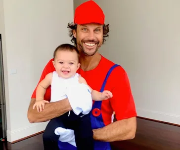 Man dressed as Mario holding a smiling baby.