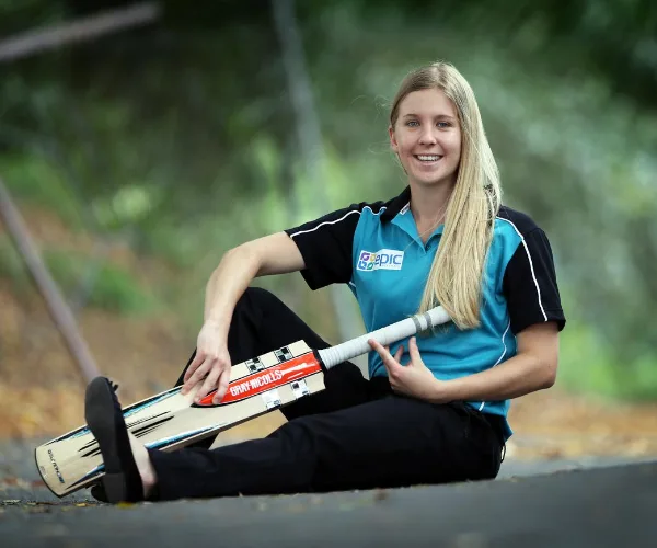 Young woman in sports attire sits on a path holding a cricket bat, smiling with greenery in the background.