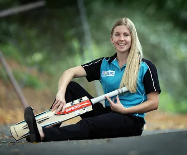Young woman in sports attire sits on a path holding a cricket bat, smiling with greenery in the background.