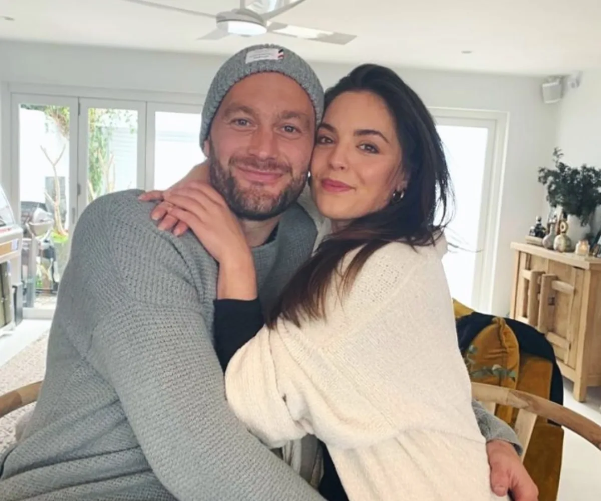 A couple, smiling and embracing, sits indoors in a cozy setting, with natural light coming through large windows.