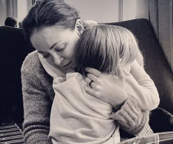 A woman lovingly embraces a young child, providing comfort and warmth, seated on a dark sofa in a cozy setting.