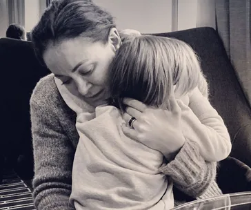 A woman lovingly embraces a young child, providing comfort and warmth, seated on a dark sofa in a cozy setting.