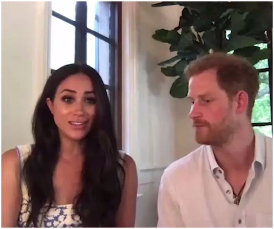 A man and woman sit together indoors, near a window, with a large plant in the background.