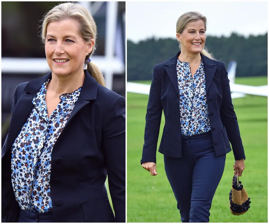 Woman in floral blouse and navy suit, smiling and walking on grass near aircraft.