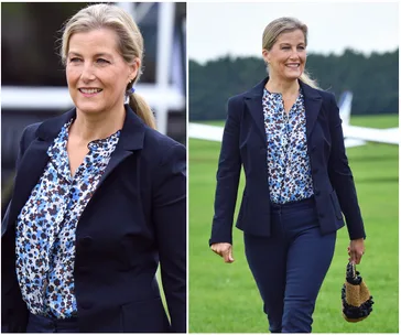 Woman in floral blouse and navy suit, smiling and walking on grass near aircraft.