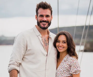 A smiling man and woman stand together outdoors, with a body of water and hills in the background.