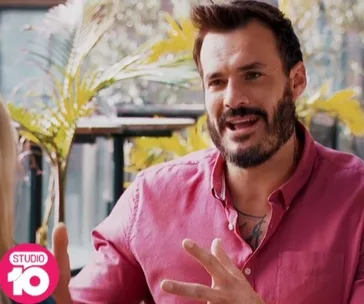 Man in a pink shirt talking energetically, with greenery in the background and a "Studio 10" logo.