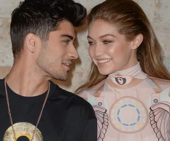 A young couple smiling at each other, the woman wearing a patterned dress and the man in a black shirt with a necklace.