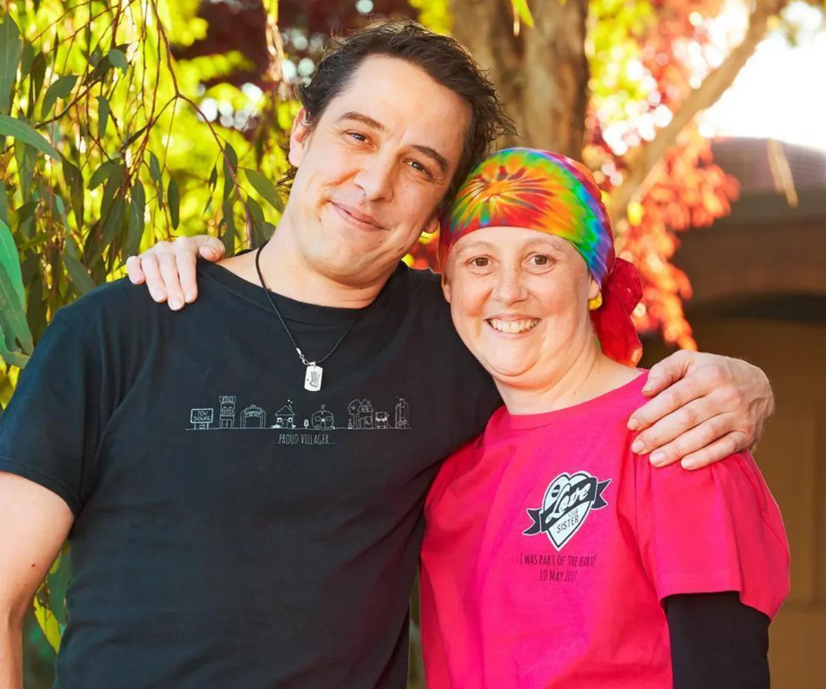 A smiling man and woman with a colorful headscarf embrace outdoors among green foliage and red leaves.