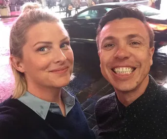 Two people smiling for a selfie on a rainy street with blurred car lights in the background.