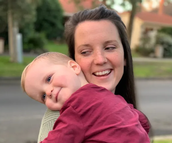 A woman smiling while holding a young child outdoors, both in casual clothing.