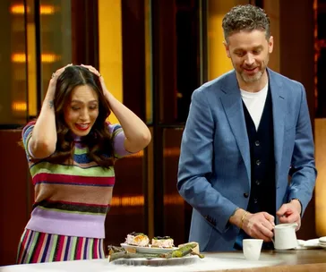 Two people react with excitement and joy at a plate of gourmet food on a cooking show set.