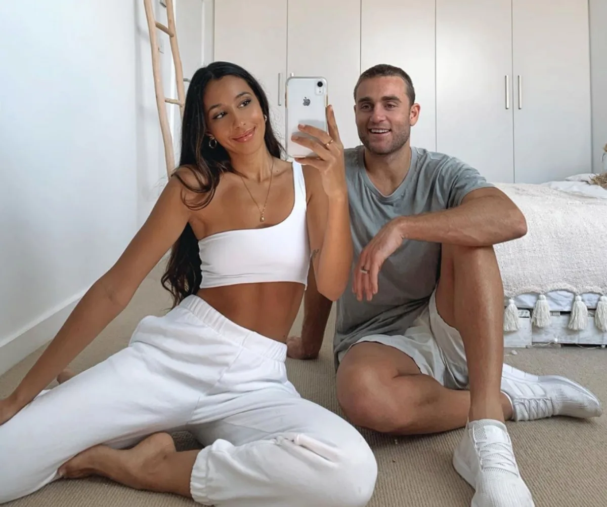 A couple in casual attire sitting on a bedroom floor, smiling and taking a selfie.