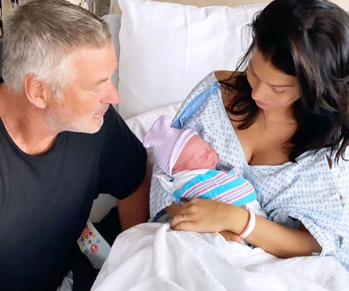 Man and woman in hospital holding newborn baby wrapped in striped blanket; woman in hospital gown.
