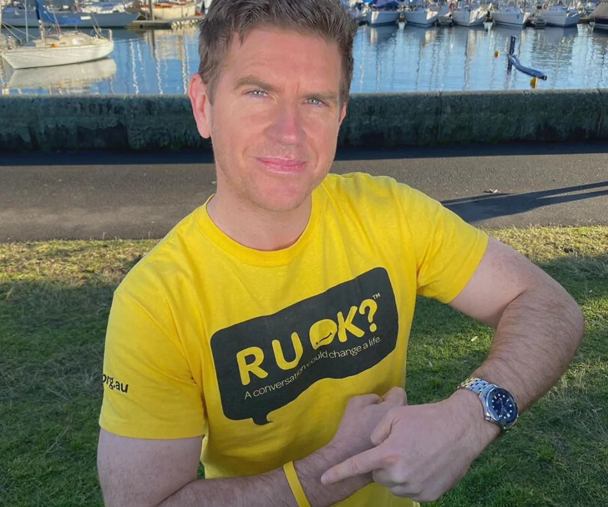 Man in a yellow "R U OK?" shirt sitting outdoors near a marina, pointing to a wristband.