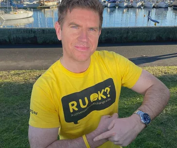 Man in a yellow "R U OK?" shirt sitting outdoors near a marina, pointing to a wristband.