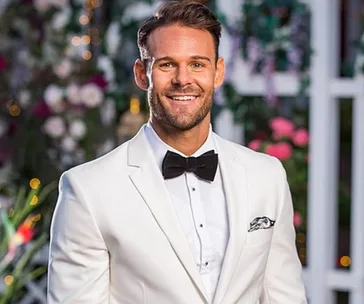 Man in a white tuxedo with a black bow tie, smiling with a floral background.