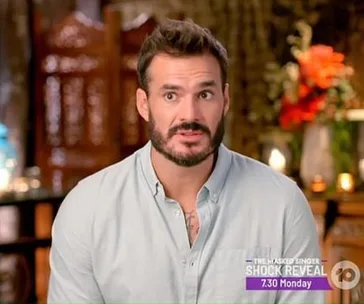 A contestant from The Bachelor Australia 2020 in a light blue shirt with a shocked expression, flowers in the background.