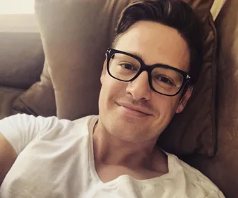 Man with glasses, wearing a white t-shirt, relaxed on a couch, smiling at the camera.