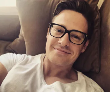 Man with glasses, wearing a white t-shirt, relaxed on a couch, smiling at the camera.