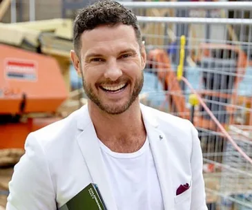 Man in a white blazer smiling at a construction site with machinery and fencing in the background.