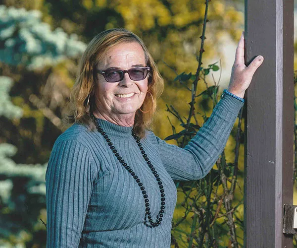 Person with long hair and glasses smiling, leaning on a wooden post, wearing a blue sweater and black bead necklace outdoors.