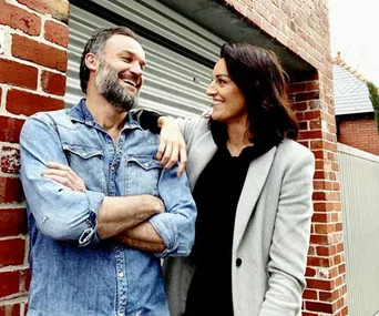 Man in denim shirt and woman in grey coat smiling and leaning against a brick wall.