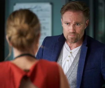 "Christian Green, a character in Home and Away, talking to a woman in a red dress with his serious expression."