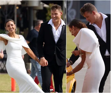 A smiling couple in wedding attire, the woman in a white dress and the man in a black suit, walking and holding hands.