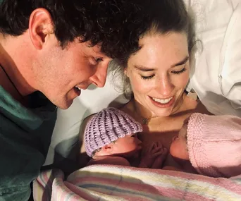 A couple lovingly gazes at newborn twins wrapped in blankets, one wearing a purple hat and the other a pink hat.