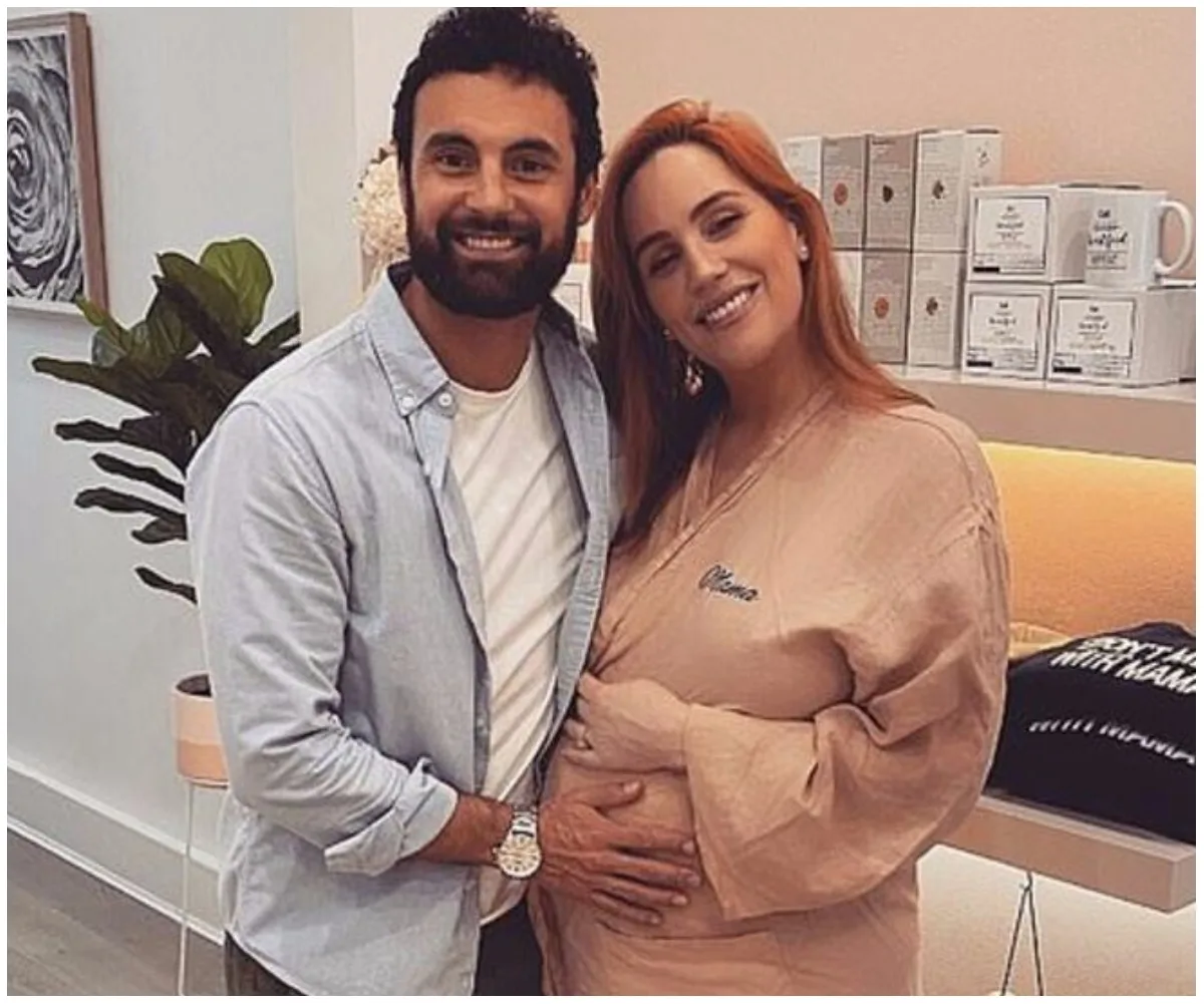 A couple smiling, with the woman holding her baby bump, standing in a cozy, well-lit room with plants and products on shelves.