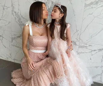 Mother and daughter in matching pink dresses and tiaras, posed against a marble wall, sharing a sweet moment.