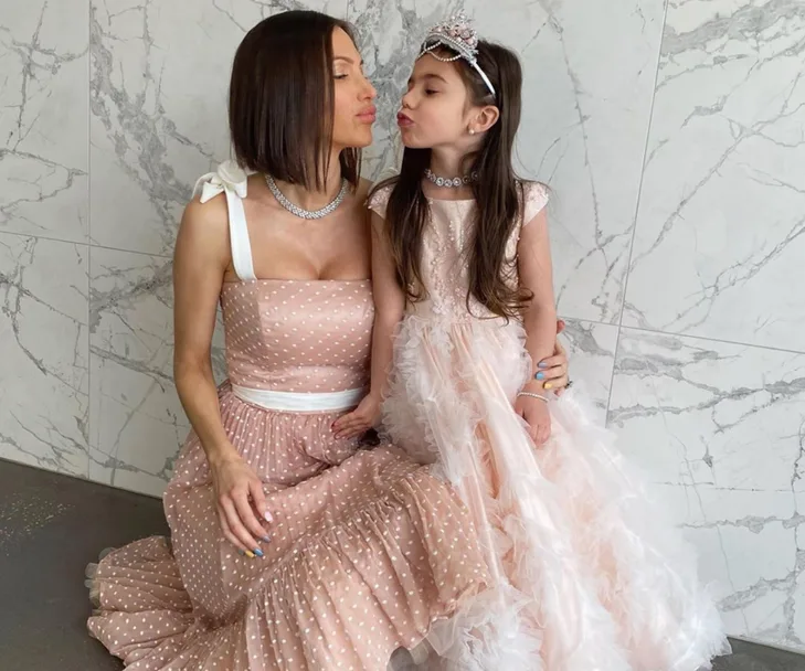 Mother and daughter in matching pink dresses and tiaras, posed against a marble wall, sharing a sweet moment.